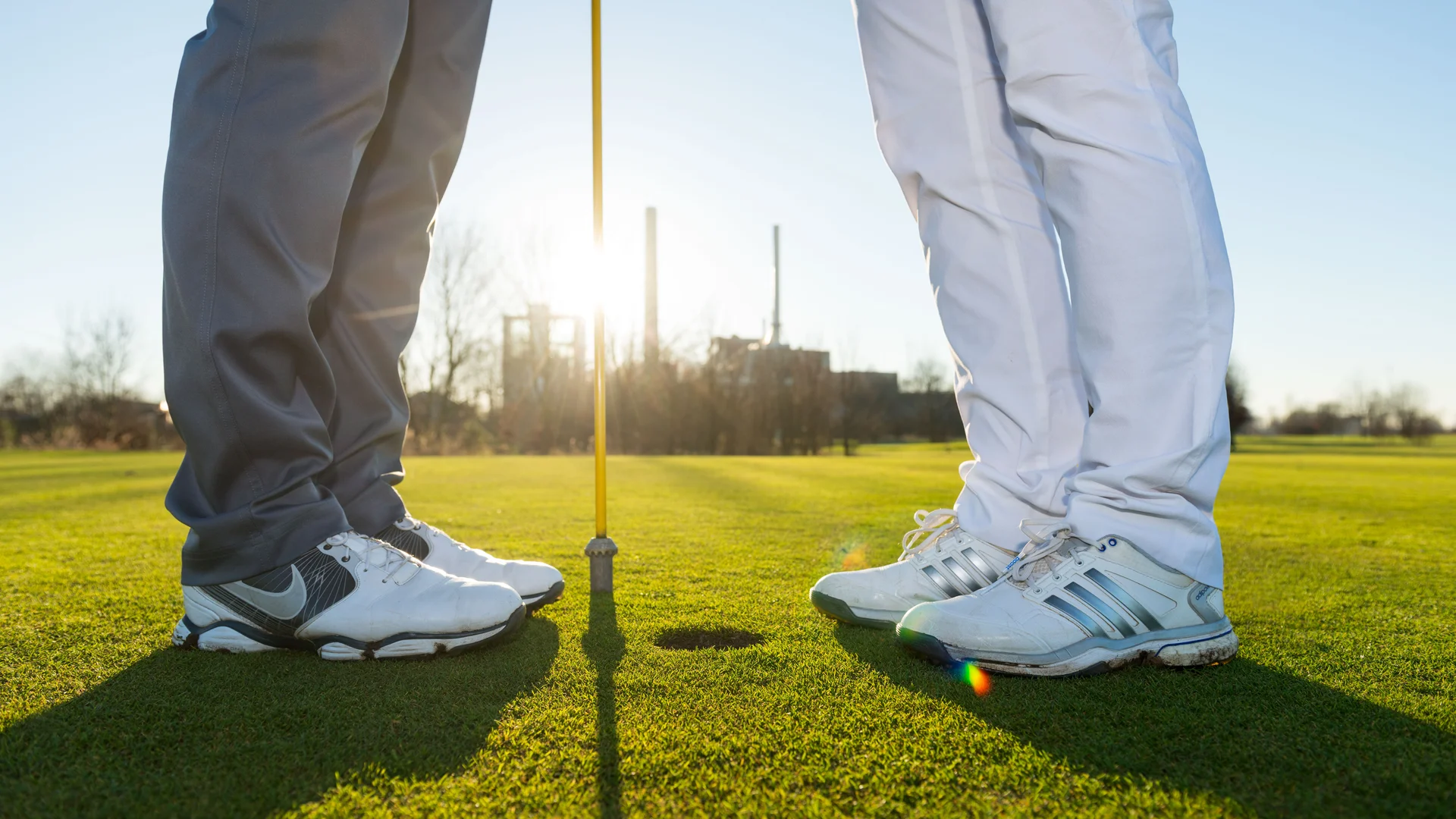 Zwei Personen in Turnschuhen stehen voreinander vor einem Golfloch mit Fahne, Ausschnitt bis kurz über dem Knie
