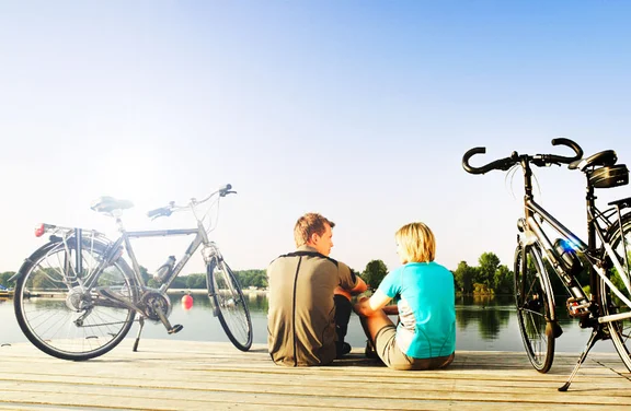 zwei Personen sitzen auf einem Steg an einem See, links und rechts jeweils ein Fahrrad, blauer Himmel