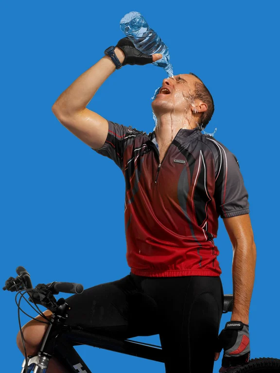 Mann in Sportkleidung auf einem Fahrrad übergießt sich mit Wasser aus einer Flasche, Blick von schräg unten
