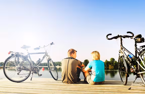 zwei Personen sitzen auf einem Steg an einem See, links und rechts jeweils ein Fahrrad, blauer Himmel