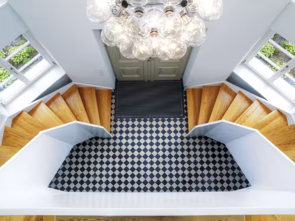 Eingangshalle von oben, karierter Fliesenboden, recht und links geht eine hellbraune Holztreppe nach unten, 