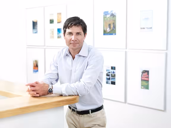 Portrait von einem Mann vor einer Wand mit gerahmten Bildern, an einem Tresen gelehnt