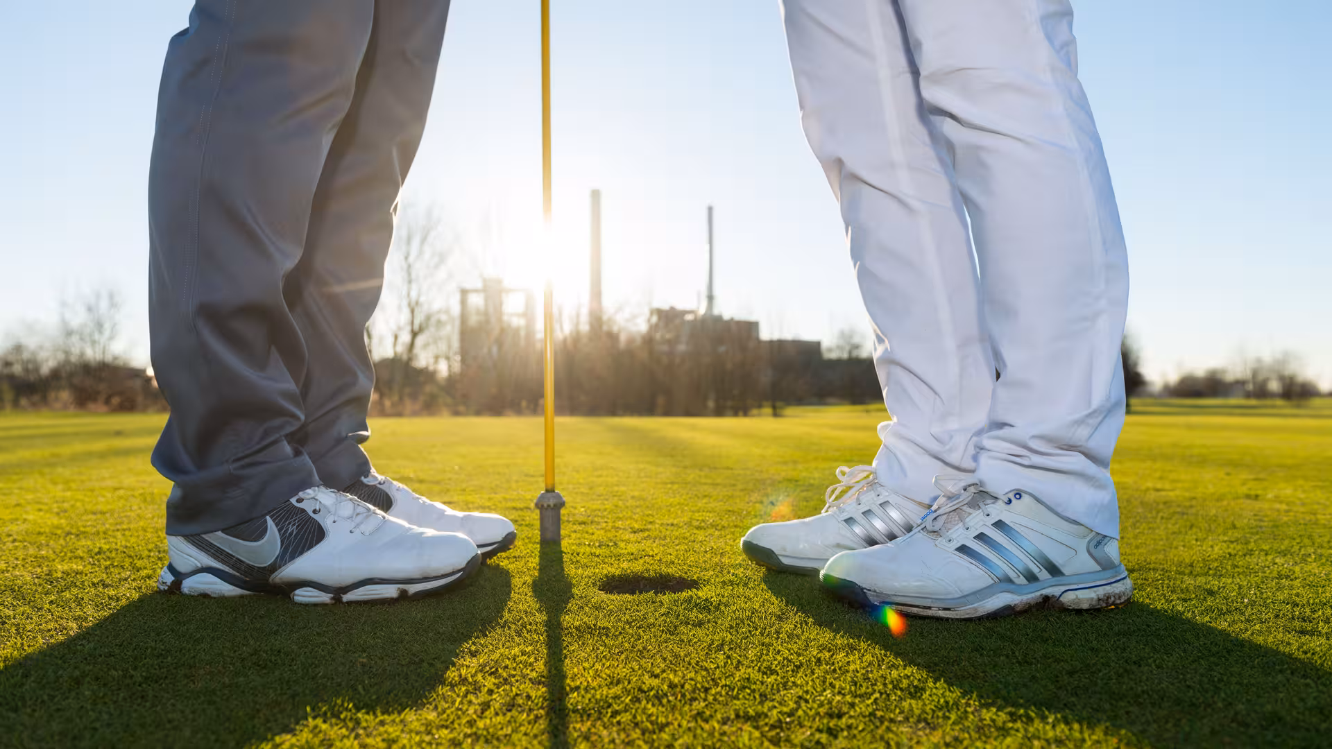 Zwei Personen in Turnschuhen stehen voreinander vor einem Golfloch mit Fahne, Ausschnitt bis kurz über dem Knie