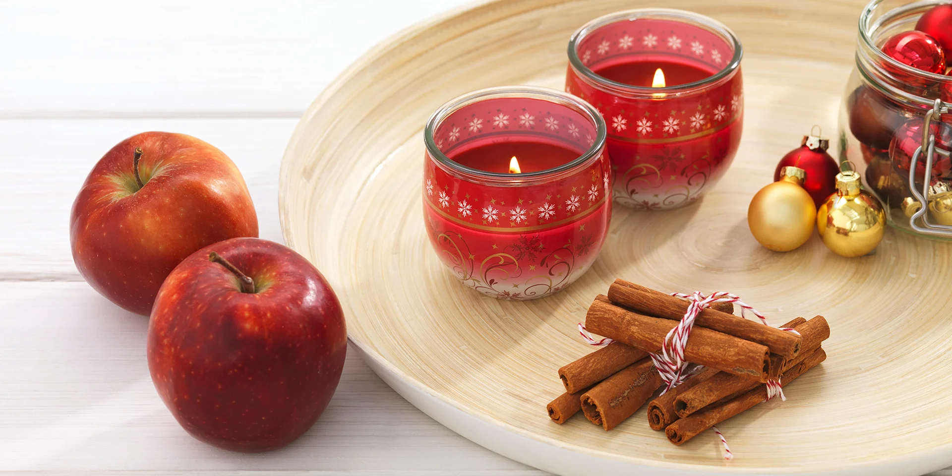Zwei Weihnachtliche Kerzen auf einer Holzschale, Zimt, kleine Weihnachtskugeln und Äpfel daneben