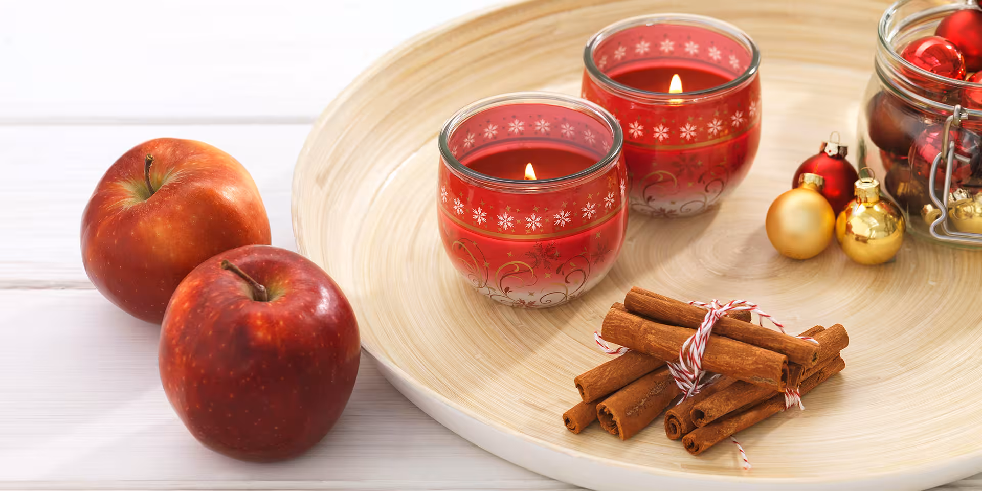Zwei Weihnachtliche Kerzen auf einer Holzschale, Zimt, kleine Weihnachtskugeln und Äpfel daneben